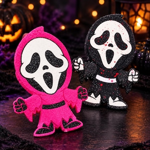 May include: Two glittery, stylized ghost figures with elongated white faces and black eye and mouth cutouts. One figure is bright pink with black accents, while the other is black with white accents and red paint splatters resembling blood.