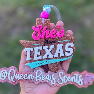 May include: A glittery, brown Texas-shaped air freshener with the words "She's from TEXAS I CAN TELL" in pink and white. The air freshener also features the text "@Queen Beas Scents" in pink glitter.