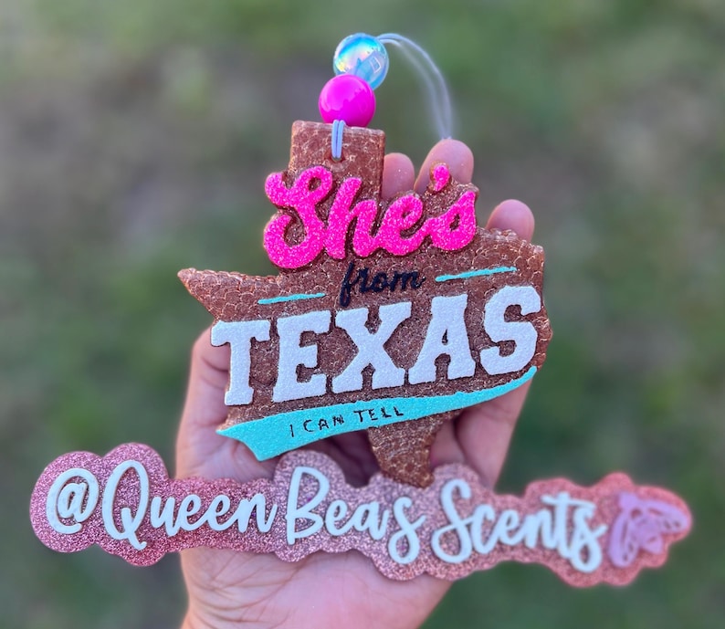 May include: A glittery, brown Texas-shaped air freshener with the words "She's from TEXAS I CAN TELL" in pink and white. The air freshener also features the text "@Queen Beas Scents" in pink glitter.