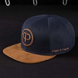 Könnte beinhalten: Marineblaue Baseballkappe mit braunem Wildleder-Schirm. Die Kappe hat ein rundes Logo auf der Vorderseite und die Worte "Pablo & Capone" auf der Seite gestickt. Ein modisches Accessoire.