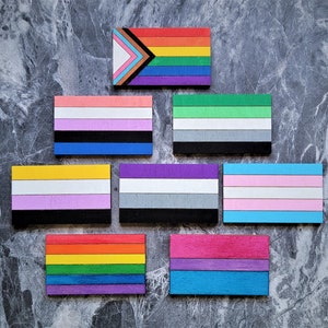 Può includere: Una collezione di otto diverse bandiere arcobaleno, ognuna con uno schema di colori e un design unici. Le bandiere sono disposte a forma di piramide su uno sfondo grigio.