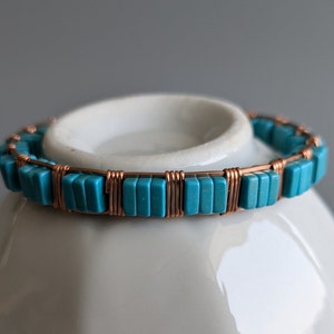 May include: A turquoise and copper bracelet. The bracelet features rectangular turquoise beads strung together with copper wire. The copper wire wraps around each bead, creating a secure and decorative design. The bracelet is displayed on a white surface.