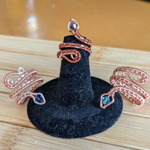 May include: Copper wire snake rings with blue and purple crystal accents. The rings are displayed on a black ring holder. The snake rings are a unique jewelry item.