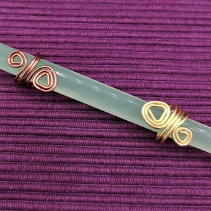May include: A light green, rectangular crystal wand with copper and gold wire wrapping. The wire is shaped into decorative spirals and triangles. The wand is set against a purple textured background.