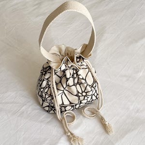 Puede incluir: Un bolso tipo saco de lona color crema con un patrón de cuadrícula floral dibujado a mano en negro. El bolso incluye una asa superior ancha y dos cordones de cuerda trenzada gruesa con extremos deshilachados para cerrar la abertura.
