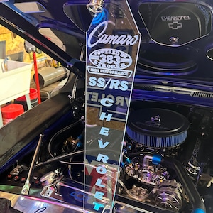 May include: A clear acrylic plaque with the text "Camaro" and "Powered by 383 Stroker High Performance SS/RS Chevrolet" in blue lettering. The plaque is attached to the engine of a blue car.