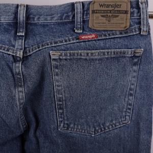 May include: Close-up of blue denim Wrangler jeans. The jeans feature a back pocket, belt loops, and a brown leather patch with the Wrangler logo. A red Wrangler tag is visible. The jeans are made of a durable denim material.