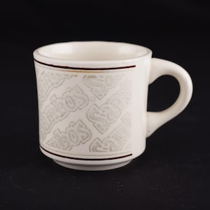 May include: Cream-colored ceramic mug with a rounded handle. The mug features the embossed text "Sambo's" in a repeating pattern around the body, framed by thin brown lines. The mug is set against a black background.