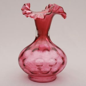 May include: A cranberry-colored glass vase with a ruffled rim. The vase has a bulbous body with a textured pattern and a slender neck. The base is a solid, rounded shape. The vase is likely vintage or antique.