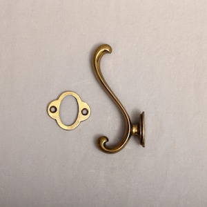 May include: A brass-colored wall hook with a curved design and a matching oval-shaped mounting plate. The hook has a decorative, scrolled shape, and the plate has two screw holes for installation. Hardware for home decor.