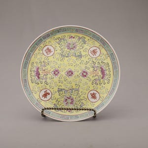 May include: A round, yellow ceramic plate with a decorative floral design and Chinese characters. The plate has a white rim and a border with green and purple accents. It is displayed on a metal stand.