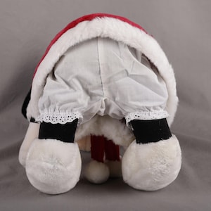 Pu&ograve; includere: Una figura di Babbo Natale in peluche, vista da dietro, con un cappello rosso e bianco rifinito con finta pelliccia. La figura indossa pantaloni bianchi con finiture in pizzo, stivali neri e grandi piedi di peluche bianchi. Le gambe rosse della figura sono visibili.