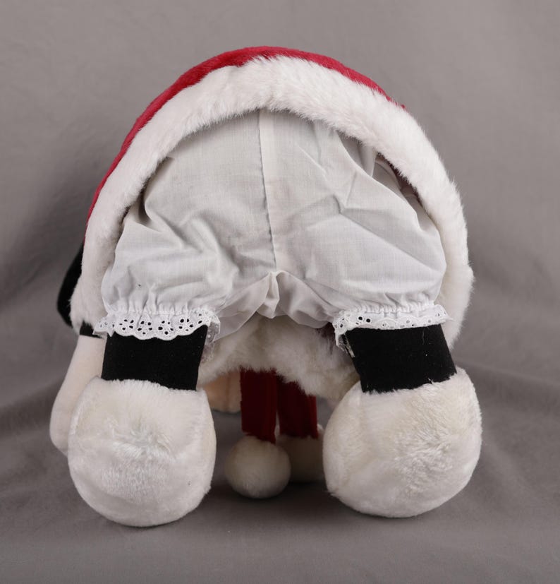 Pu&ograve; includere: Una figura di Babbo Natale in peluche, vista da dietro, con un cappello rosso e bianco rifinito con finta pelliccia. La figura indossa pantaloni bianchi con finiture in pizzo, stivali neri e grandi piedi di peluche bianchi. Le gambe rosse della figura sono visibili.