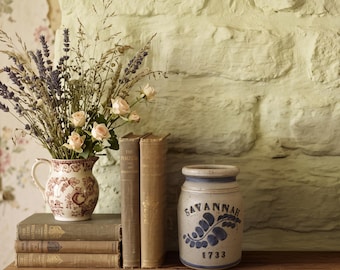 May include: A ceramic pitcher with a red floral pattern filled with dried lavender and pale pink roses, placed on a stack of three vintage books next to a beige stoneware crock featuring the blue text SAVANNAH 1733 and a fern leaf design.