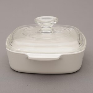 May include: A white, square casserole dish with a clear glass lid and a rounded glass handle. The dish has two small handles on the sides. The dish is on a gray background.