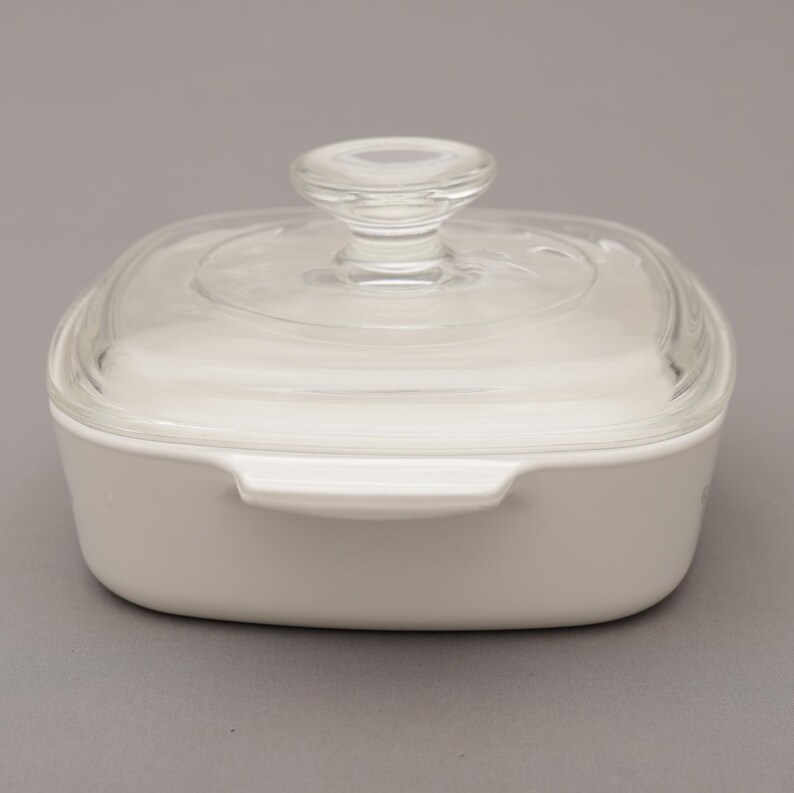May include: A white, square casserole dish with a clear glass lid and a rounded glass handle. The dish has two small handles on the sides. The dish is on a gray background.