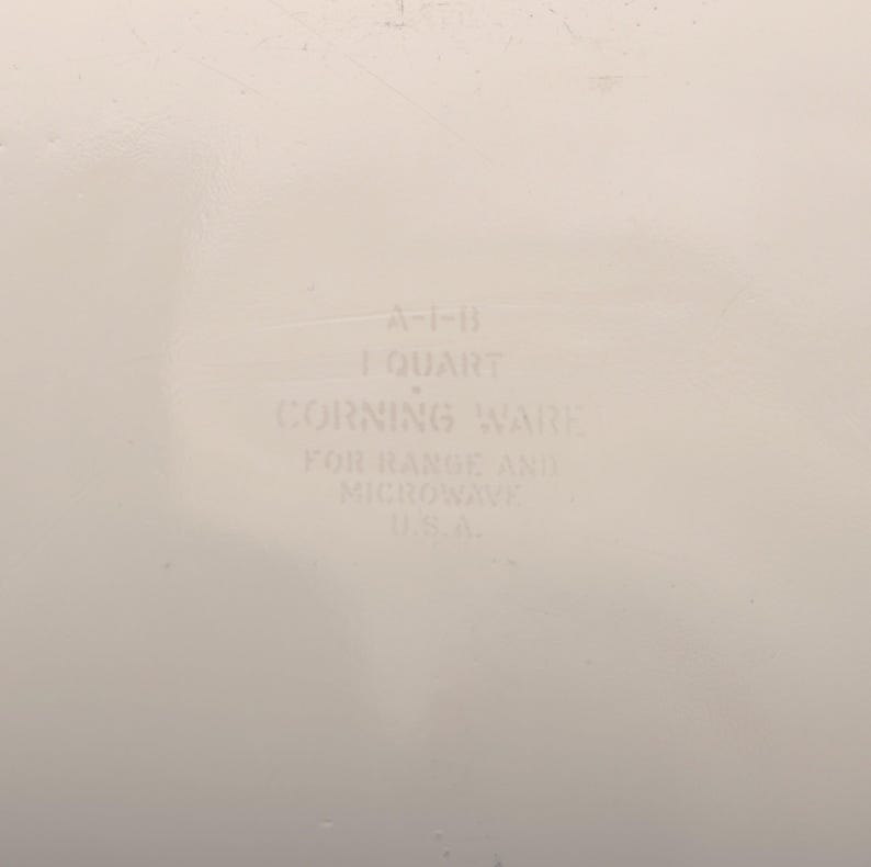 May include: Off-white Corning Ware dish with text that reads "A-1-B 1 QUART CORNING WARE FOR RANGE AND MICROWAVE U.S.A.". The dish is designed for use in both the range and microwave.