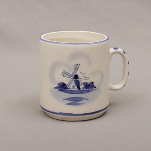 May include: A ceramic mug with a cream-colored base and a blue rim and handle. The mug features a blue illustration of a windmill and water scene. The handle has blue polka dots.