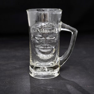 May include: A clear glass beer mug featuring a molded relief of a person's face on the front surface. The base of the mug displays the embossed signature text Joe Namath. The vessel includes a sturdy handle on the right side and a smooth rim.