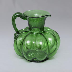 May include: A translucent emerald green glass pitcher featuring a rounded, pumpkin-like body with vertical ribbed indentations. The vessel has a wide, flared rim, a curved handle attached to the side, and a small pouring spout at the top.