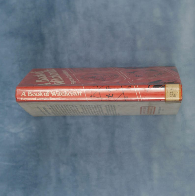 Puede incluir: Un libro titulado "A Book of Witchcraft" de Raymond Lamont Brown. El libro tiene una cubierta roja con texto blanco y un lomo blanco. El libro est&aacute; sobre una superficie azul.