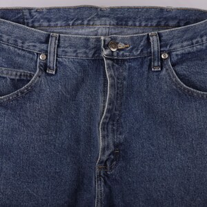 May include: Close-up of blue denim jeans. The image shows the front of the jeans, including the button closure, belt loops, and front pockets. The denim has a classic, slightly faded appearance.