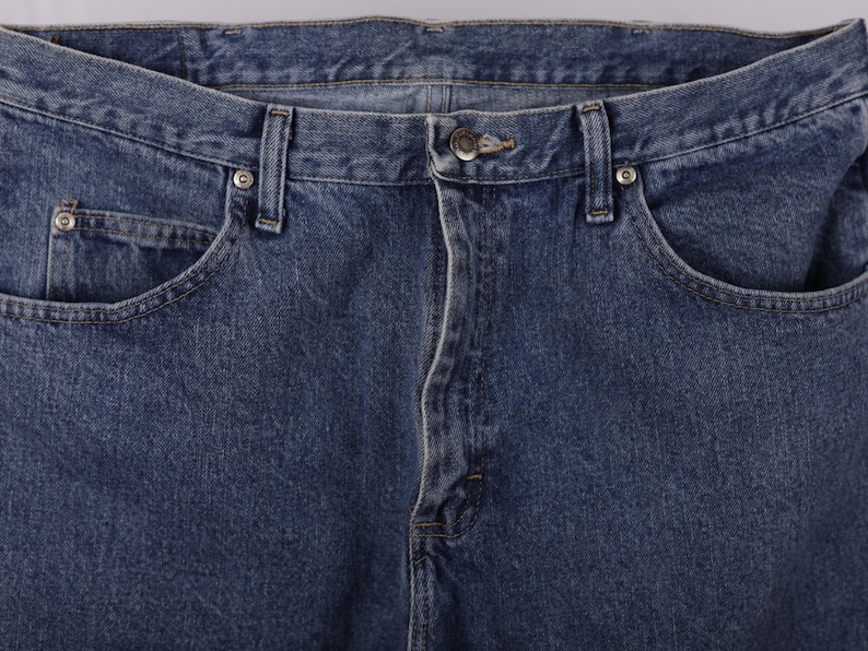 May include: Close-up of blue denim jeans. The image shows the front of the jeans, including the button closure, belt loops, and front pockets. The denim has a classic, slightly faded appearance.