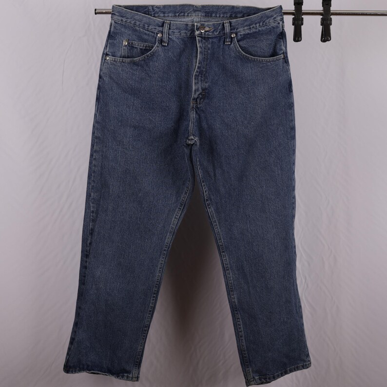 May include: Blue denim jeans with a classic five-pocket design. The jeans are hanging on a metal rack. The jeans have a straight leg cut and a button and zip closure. The jeans are made of a durable denim material.
