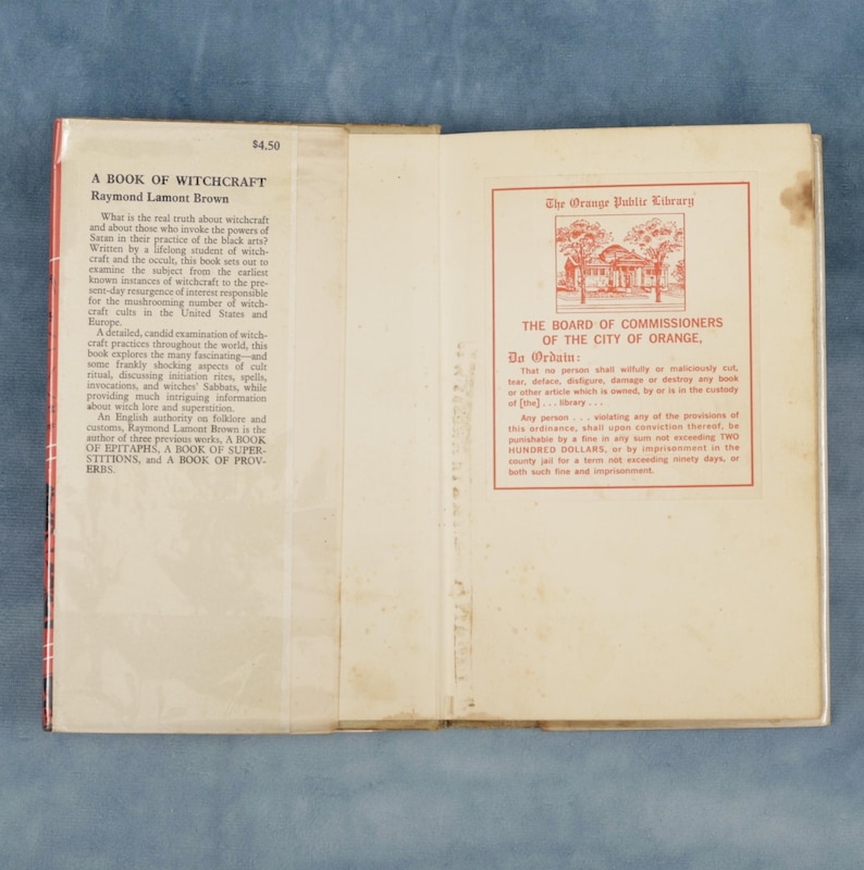 Puede incluir: Libro abierto titulado "A Book of Witchcraft" de Raymond Lamont Brown. La portada es roja y blanca con texto. La p&aacute;gina derecha tiene un sello rojo de "The Orange Public Library".