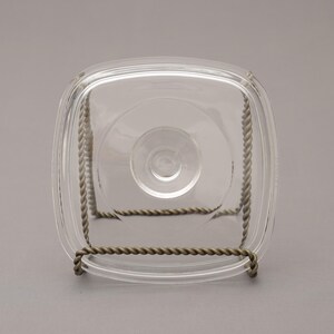 May include: A clear, square glass dish with rounded corners, resting on a decorative metal stand. The stand is made of twisted metal, providing support. The dish has a central circular indentation.