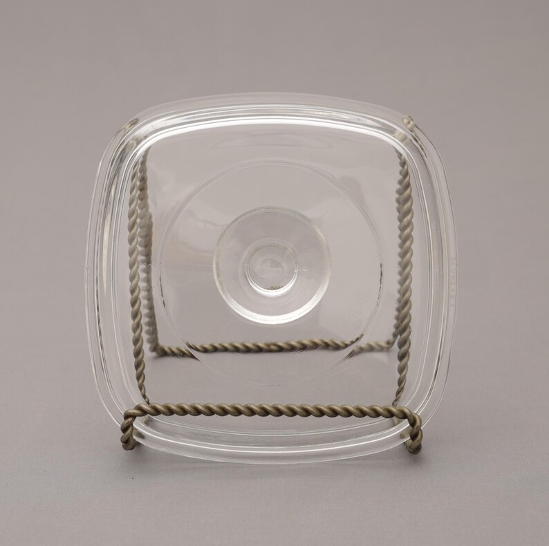 May include: A clear, square glass dish with rounded corners, resting on a decorative metal stand. The stand is made of twisted metal, providing support. The dish has a central circular indentation.