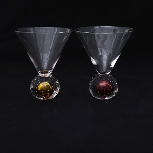 May include: Two clear glass martini glasses with wide, conical bowls. Each glass features a spherical base containing air bubbles and a solid colored core, one with a yellow center and the other with a deep red center, set against a black background.