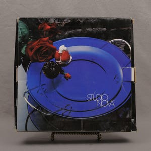 May include: A box featuring a blue plate design with the text "STUDIO NOVA". The box art includes red roses and other floral elements. The box is displayed on a decorative metal stand.
