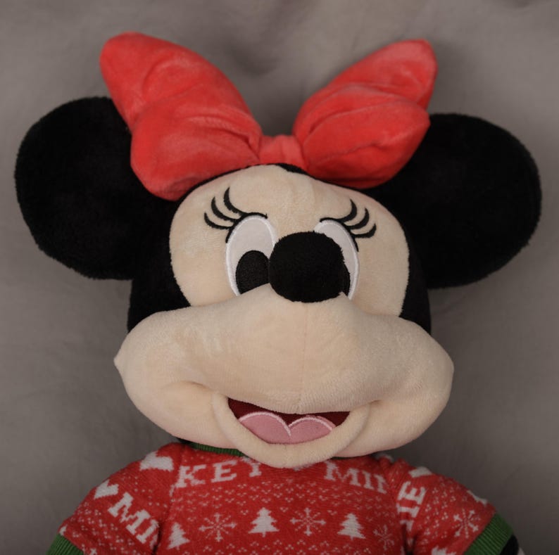 May include: A plush Minnie Mouse doll with a red bow, black ears, and a red sweater with white Christmas tree designs. The sweater has the text "KEY MINNIE" on it. The doll has a smiling expression.