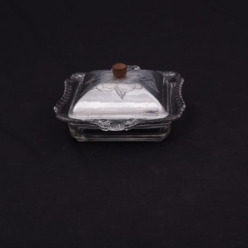 May include: A clear glass butter dish with a silver-plated lid and a wooden knob. The lid has a floral design etched into it.