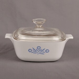May include: White square casserole dish with a clear glass lid. The dish has two handles and a blue floral design on the front. A clear glass knob is on top of the lid. The dish is likely made of ceramic or glass.
