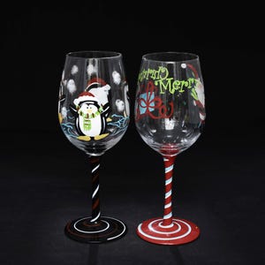 May include: Two clear glass wine glasses with painted designs. The left glass features a penguin wearing a Santa hat and scarf with white snowflakes. The right glass displays the text Merry Christmas in green with a red bow and candy cane stem.