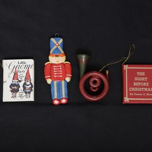 May include: Four hanging ornaments: a white book titled Little Gnome Facts with two gnome illustrations, a red and blue soldier figure, a dark red circular horn instrument, and a red square book titled The Night Before Christmas by Clement C. Moore.
