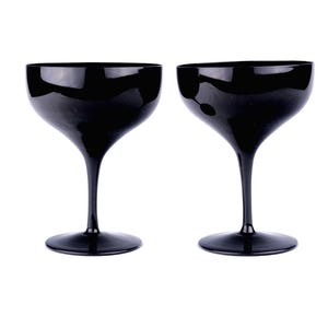 May include: Two black champagne coupe glasses. The glasses have a wide, shallow bowl and a long, slender stem. The base of the glass is round. The glasses are made of a shiny, reflective material.