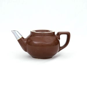 May include: A small, brown ceramic teapot with a silver spout and a handle. The teapot has a white rim around the opening and sits on a white surface. The teapot is designed for brewing tea.