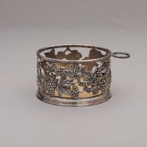 May include: Silver-toned metal wine coaster with a handle. The coaster features a detailed design of grape clusters and leaves around the sides. The interior has a gold-colored lining, and the coaster sits on a circular base.