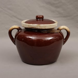May include: A brown ceramic crock with a lid. The crock has two handles and a lid with a small knob. The lid has a cream-colored rim. The crock is a deep brown color with a glossy finish. The handles have a cream-colored accent.
