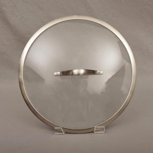 May include: A round, clear glass lid with a stainless steel rim and handle. The lid is designed for cookware and sits on a clear plastic stand. The handle is a curved, brushed metal.