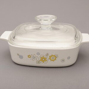 May include: A white square casserole dish with a clear glass lid. The dish features a floral pattern in yellow, gray, and blue. It has two white handles on either side, and a clear glass knob on the lid.