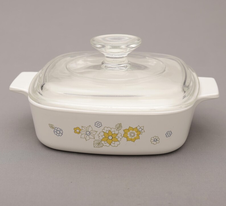 May include: A white square casserole dish with a clear glass lid. The dish features a floral pattern in yellow, gray, and blue. It has two white handles on either side, and a clear glass knob on the lid.