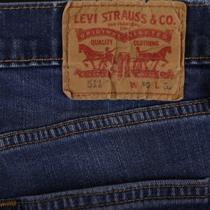 May include: Close-up of blue denim jeans with a brown leather Levi Strauss & Co. label. The label features red text and two horse-drawn carriages. The jeans have visible stitching and the label reads "511 W 30 L 32".