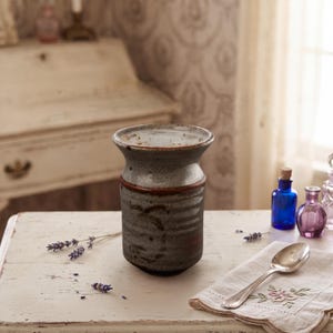 May include: A cylindrical ceramic vase with a flared rim, featuring a mottled grey and brown glaze with horizontal ribbed textures. The vase has a dark brown band around the upper section and a smooth, light-colored interior finish.