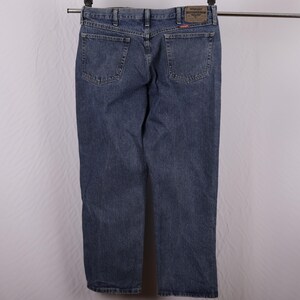 May include: Blue denim jeans hanging on a metal bar. The jeans have two back pockets and a brown leather patch with the brand name. The denim is a medium wash with some fading.
