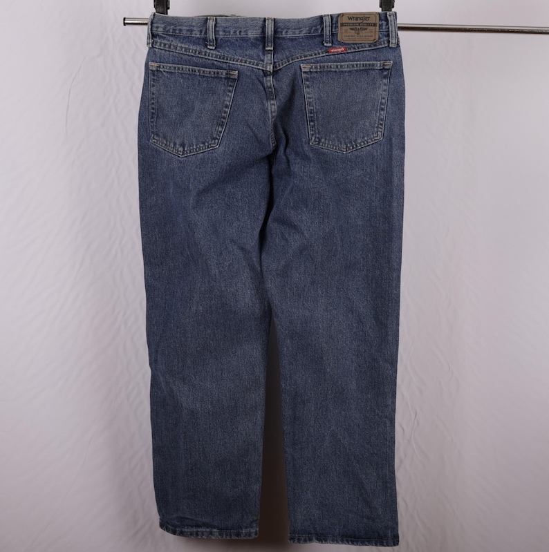 May include: Blue denim jeans hanging on a metal bar. The jeans have two back pockets and a brown leather patch with the brand name. The denim is a medium wash with some fading.