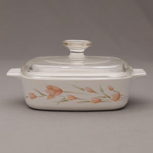 May include: A white rectangular casserole dish with a clear glass lid and a clear handle. The dish is decorated with a floral pattern in shades of peach and green. The dish has two handles on either side.